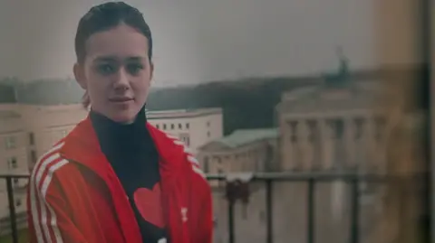Brunette white teenage girl standing on a balcony. Background is blurred buildings. She is wearing a red sports jacket and black polo neck top.