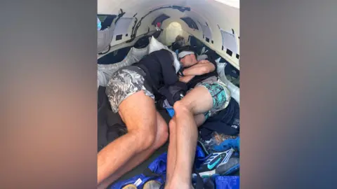 Rowmads Two men sleeping next to each other on a boat. One man is wearing a white eye mask and the other man has his head on a pillow.