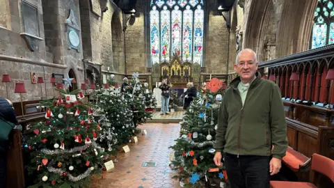 A man with grey hair and a green zipped top standing in a church with rows of decorated trees behind him and further back a large stained glass window