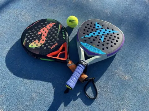 A pair of padel bats lay on the blue tarmacked ground. Between them is a green tennis ball. The racquets are similar to those used in tennis, but have a solid centre.