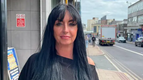 Tom MacDougall/BBC Nicola Baldwin, has long, black hair and a fringe and is standing next to Gateshead's high street outside the front of her hair salon.