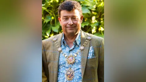 A man, Chris Aldred, smiles at the camera wearing a very decorative gold chain.