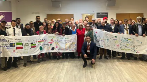 BBC/Julia Moore Group of people holding up a long white piece of silk with drawings and words on to describe their feelings on integration and race equity. The banner includes different languages and flags in many colours.