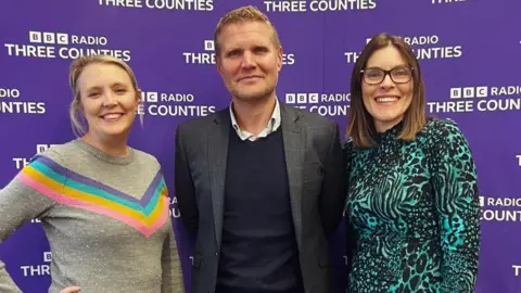 Laura, JVS and Liz stand together in front a purple BBC Three Counties banner stand. He wears a suit jacket over a v-neck jumper and white shirt. Liz wears a patterned black and turquoise top and black glasses, while Laura wears a grey jumper with a V-shape in rainbow stripes.