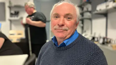 A man with short white hair and a white moustache, wearing a dark jumper and blue shirt, smiles as he sits in a room. Other people can be seen behind him.