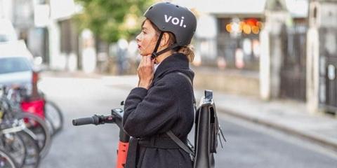 Woman in dark top standing on a scooter fastens a helmet
