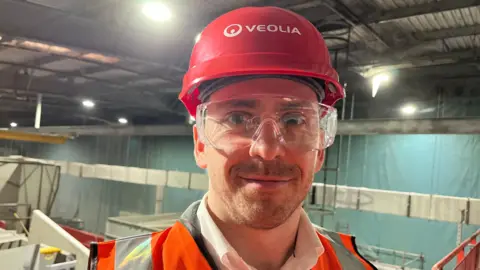 Oliver Peat standing in a large warehouse wearing safety glasses, a high-viz top and a red helmet with Veolia written on it