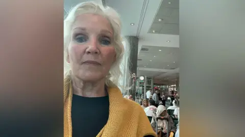 Lenise Blom Lenise Blom, who is wearing a yellow jumper and black shirt takes a selfie while stood inside the terminal at Dubai Airport. Dozens of people are sat behind her. She is a woman in her 70s who has wavy white hair.