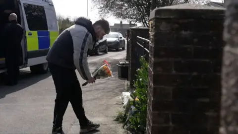 Man in a grey and black puffer jacket places flowers next to a wall