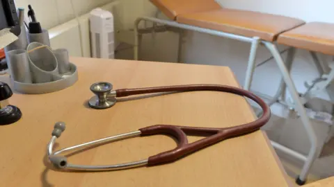 A brown-red stethoscope on a desk in a doctor's surgery. A medical bed is in the background.