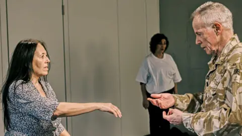 Colin Morris Photography A rehearsal scene from the dance group where a woman with long dark hair stretches her arm towards a man dressed in a soldier uniform. His hands are open and about to reach out to her. There is another woman in the background.
