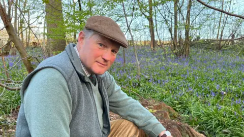 Hole Park owner Edward Barham sat in wood surrounded by bluebells in bloom
