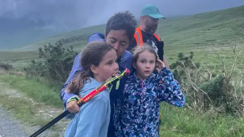 Zoe Murphy Trish Patterson hugging her twin girls and giving one of them a kiss on the head as she walks along a wild path with a man in high vis in support behind her.