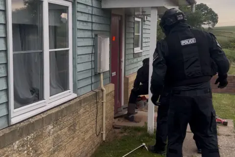 West Mercia Police Two police officers in protective uniform are visible in a shot taken during a police raid on the outside of a house. The home is built of light coloured stone bricks and pale blue wooden cladding.