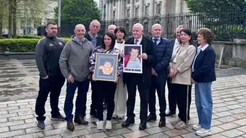 BBC A group of men and women standing together. Two of them are holding up images of two men. 