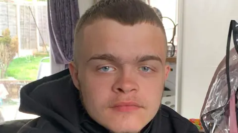A teenage boy in a black hoodie, sat on a sofa inside a living room. He is looking at the camera.