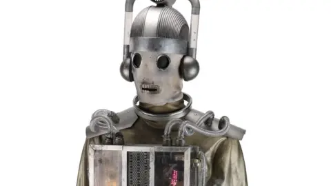 Close dari seorang cyberman Monda seorang Mondasian yang terlihat samar -samar steampunk dengan bahan seperti wajah. Ini memiliki headpiece perak besar dan kotak cahaya di dadanya