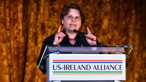 Getty Images A woman with short curly brown hair, wearing glasses and a black shirt, stood in front of a pulpit that reads US-Ireland Alliance, she has both her index fingers pointing upwards as she speaks into a microphone