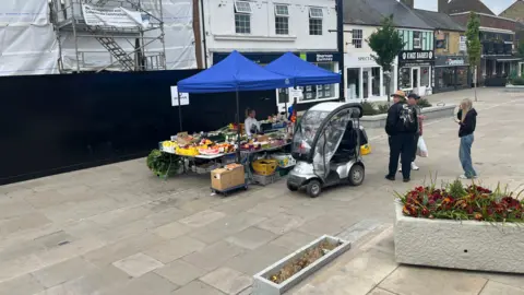 John Devine/BBC Two small blue gazebos side by side with tables beneath with fruit and veg displayed, a silver mobility scooter with some people standing around it is in front of the stall, with shops behind and a building on the left being demolished with black fencing around it. There are a couple of planters in the foreground.