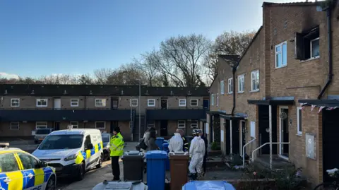 Luke Deal/BBC Two police cars parked outside a block of terrace homes in Ipswich. A police officer and forensics specialists are standing in front of the entrance to one of the homes and next two three bins.