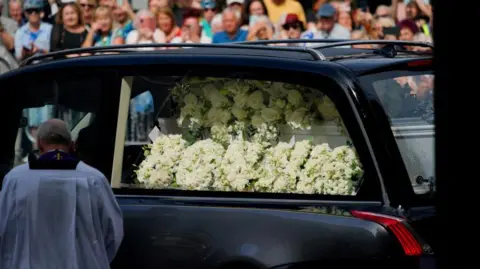 PA Media A floral tribute with white flowers spells out 'Bonnie'. White roses and lilies are placed on her coffin inside the hearse as it arrives at the church. A priest can be seen standing next to the hearse with his head bowed. Blurred out crowds of people can be seen behind the hearse.