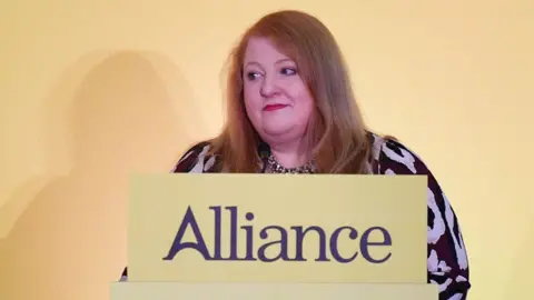 Naomi Long, a woman with shoulder-length ginger hair and red lipstick, is standing in front of a yellow podium which is topped with a sign showing the Alliance Party logo.  She is wearing a black and taupe patterned dress and a chunky necklace. 