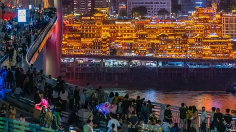 NurPhoto via Getty Images People crowding on a bridge overlooking a cluster of traditional-looking Chinese buildings, glowing in golden light.