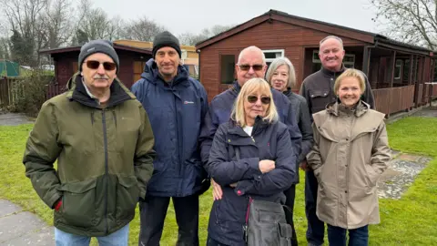Four men and three women are standing together in a line and looking at the camera. They are wearing winter coats and standing next to a former bowling green with two wooden cabins. A woman at the front has blonde hair and wears sunglasses and has her arms crossed.
