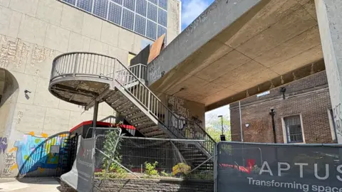 A concrete staircase with metal railings has a twist as it rises to a concrete bridge which is dotted with graffiti. The staircase is cordoned off by metal fencing. There is a tall concrete building, similarly tagged, to the left and an older dark brick building with dark greay windows behind the bridge, of which only the ground floor is visible. On one of the temporary fences, a banner shows the logo of a company that claims to be "Transforming spaces". 