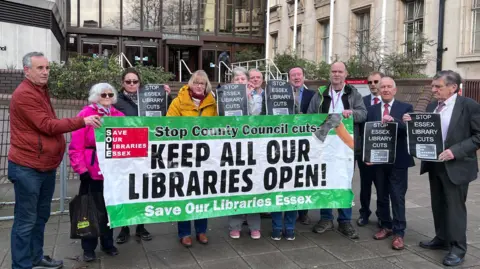 About a dozen people standing outside an office building. Many of them are holding signs saying "STOP ESSEX LIBRARY CUTS" in white lettering on a black background. In front of them is a white and green banner that says: "Stop County Council cuts- KEEP ALL OUR LIBRARIES OPEN! Save Our Libraries Essex."