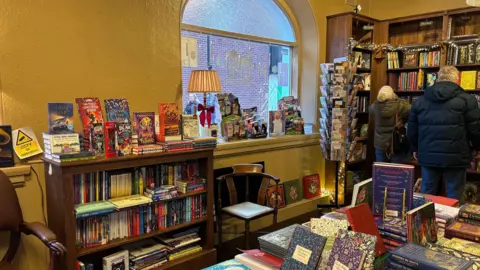 Inside a bookshop with a large window looking out onto the street. There are dark brown wood bookshelves filled with books, and fairy lights draped over them. A table in the middle of the room is stacked with books and there are brown wooden chairs next to the shelves. Two customers browse in the back of the room with their backs to the camera