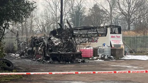 Two burnt out buses, one is black metal melted parts and another still has a partial boyd frame, it is a white bus with pink writing that says Allen's Tours. There is other debris at the buses and a police cordoned line with white and red tape - in red it says DO NOT CROSS and POLICE CORDON 