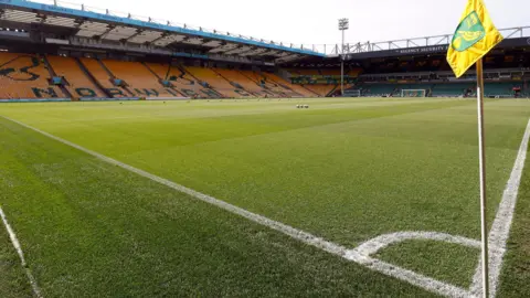 PA Media Norwich City's Carrow Road ground showing the football pitch with white markings on it. In the background is one of the stands which has yellow and green seating spelling out "Norwich City". In the foreground is a corner post with the Norwich City yellow and green flag at the top.