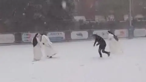 Three nuns in white clothing and black headdress in a snowball fight with a priest dressed in black.