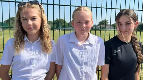 Three young girls, aged around 12-14, standing in a line in front of metal railings with a football pitch in the background. The girl on the left has long dark blonde hair with sunglasses perched on top of her head and is wearing a white blouse. The girl in the middle is also wearing a white blouse and has strawberry blonde hair tied back in a centre parting. The girl on the right has long dark hair in pigtails and is wearing a black Nike t-shirt.