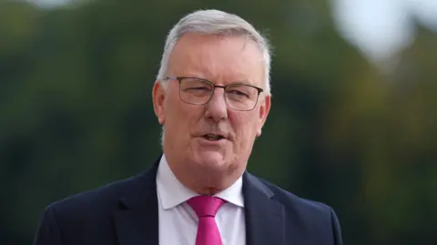 PA Media A man is talking to someone off-camera. He has white hair and is wearing dark rimmed glasses. He is wearing a dark-colours suit jacket, a white shirt and a pink tie. The background is out-of-focus, but there is an outline of greenery. 