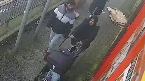 West Yorkshire Police Man in striped puffa jacket stood next to a woman in a dark headscarf pushing a pram on a narrow pavement