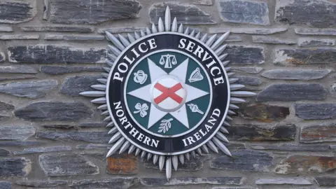 A green and black psni badge on a grey stone wall