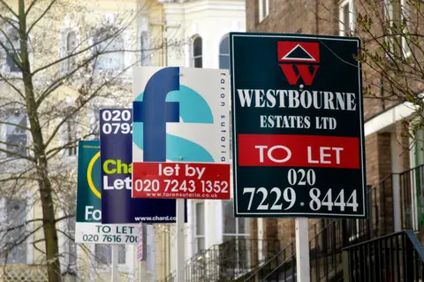 Four different "to let" signs from different estate agents standing in a street of terraced houses