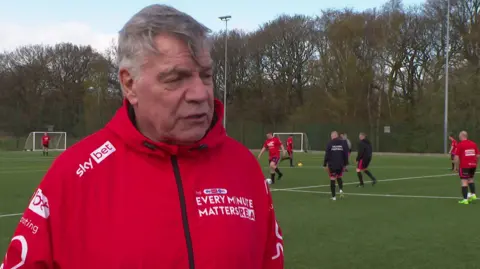 A man with short grey hair and wearing a red jacket with 'Every Minute Matters' branding is standing in front of a football pitch. Several men are playing football behind him.