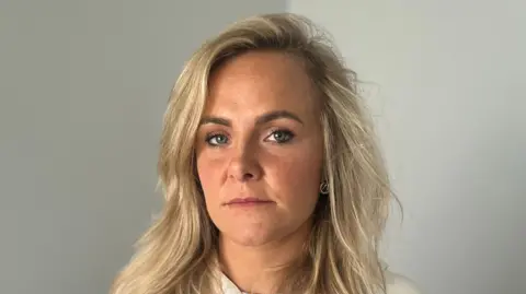 Hannah Farrell, a nurse, looks into the camera. She has a neutral expression on her face. We can see her from the shoulders up. She has shoulder-length blonde hair and is wearing a white or cream top. She has a metal chain around her neck. She is standing in a room, there is a plain grey-coloured wall behind her.