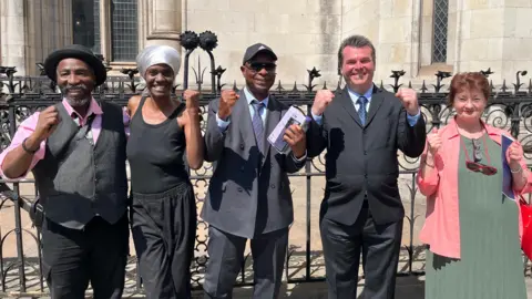 Campaigners pose with raised fists outside a court building after a legal victory. The group stand in front of ornate railings and stone Gothic-style architecture, with one person holding court documents.