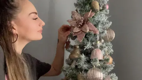 Sally-Anne Long Sally-Anne Long, a woman with her hair tied back wearing a grey t-shirt, looks at a Christmas tree decorated with gold and red ornaments.