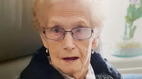 Ellen Watson wears a dark woollen coat with a white and grey scarf and a brooch. The sunlight from the window behind her is shining through her short blonde hair. She is wearing dark-rimmed glasses. 