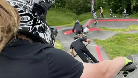 A biker is visible in the foreground, with a helmet on. In the background a track is visible with humps and slopes, with cyclists riding up and down them. Many of those pictured are young people. 