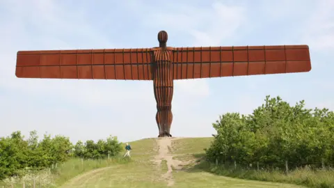 PA Media The Angel of the North is a huge copper-coloured sculpture standing atop a hill. It has a human body - with arms replaced by giant wings.