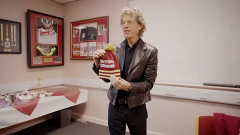 Pete Bresser Sir Mick Jagger wearing a dark shirt and leather jacket, holding a burgundy bobble hat with a yellow bobble and white stripes at the bottom. Text on the hat reads Dartford Grammar. Framed sports memorabilia is on the wall behind him and there is a china tea set laid out on a table with white and burgundy tablecloths.