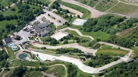 A birds eye view looking down on a dry ski slope snaking through a grass area with trees and buildings