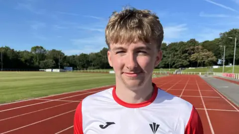 Close up picture of Ewan smiling looking at camera. His eyes are squinting due to the sun. Behind him is an athletics running track with trees in the background. Ewan wearing a white and red top and has blonde hair.