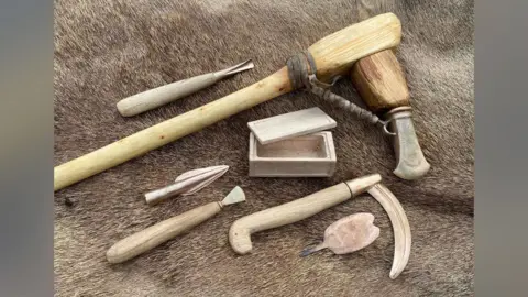 EMMA JONES/ANCIENT CRAFTS Four wooden handled replica Bronze Age tools, plus a replica Bronze Age box, arrow head and blade against a fur background